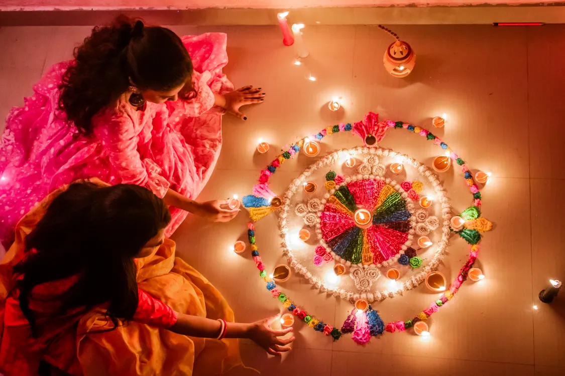 Indian women arranging glowing diyas around a colorful rangoli for Diwali